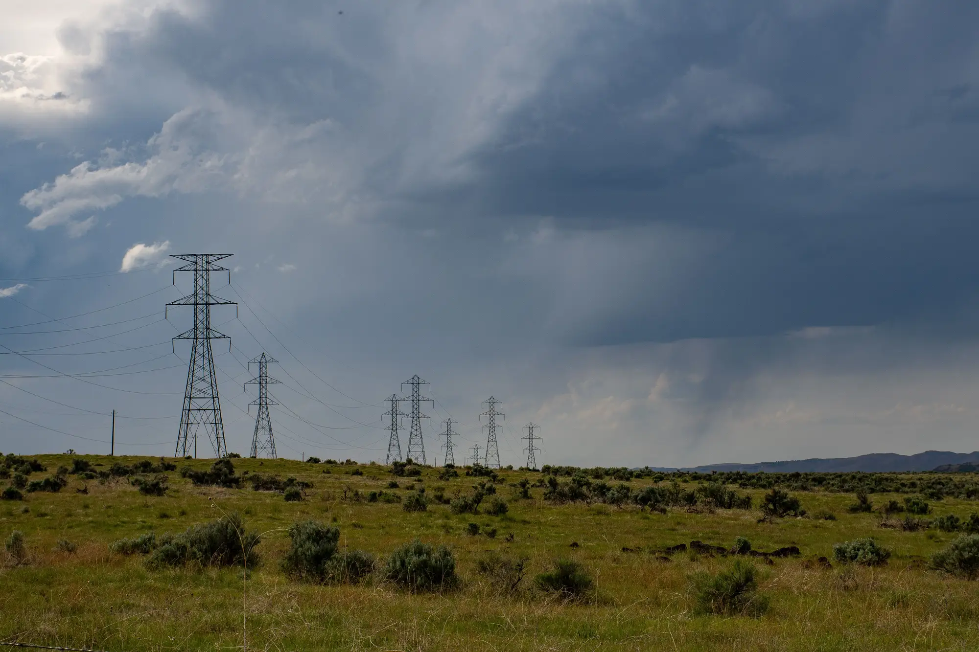 A line of steel lattice transmission towers running across sagebrush range under a rain-heavy sky